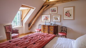 Un dormitorio con techo inclinado, una cama con colcha roja, un aparador, dos sillas y cuadros con motivos florales en la pared.