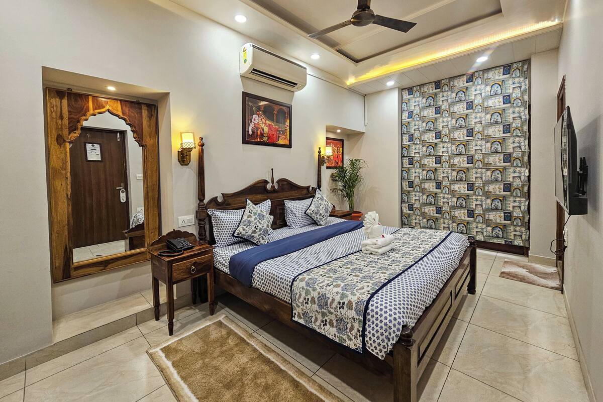 A bedroom with a wooden bed, a nightstand, a ceiling fan, an air conditioning unit, a wall-mounted TV, and a patterned curtain.