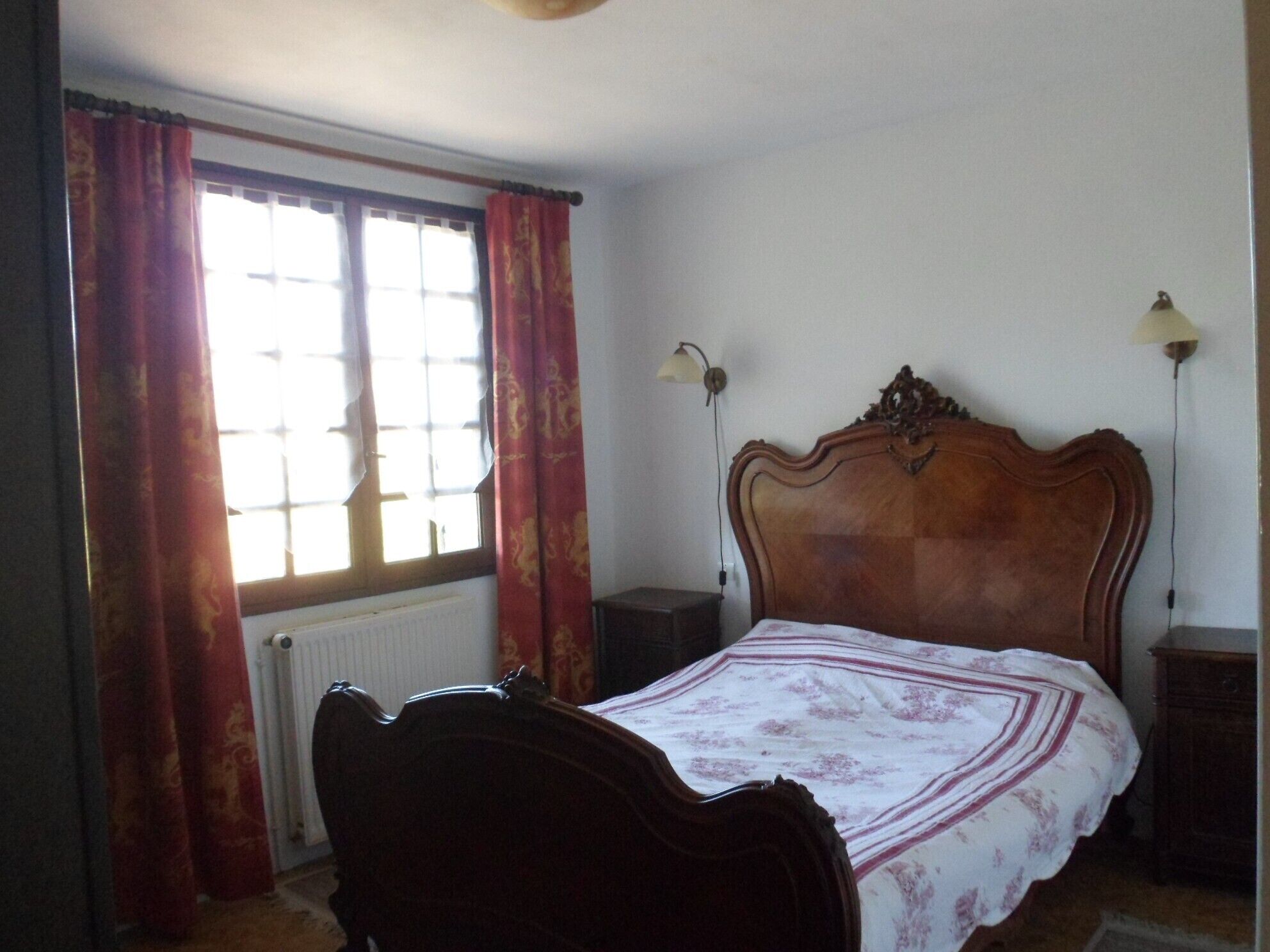 Un dormitorio con una cama de madera, un radiador, una ventana con cortinas y dos lámparas fijadas a la pared.