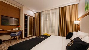 A hotel room with a bed, a desk, a chair, a TV, and a window with curtains. - Junior King Comfort Sunday Emerald (Jeddah)