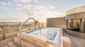En jacuzzi på taket med en glasräcke, ett trädäck och en modern byggnad i bakgrunden.