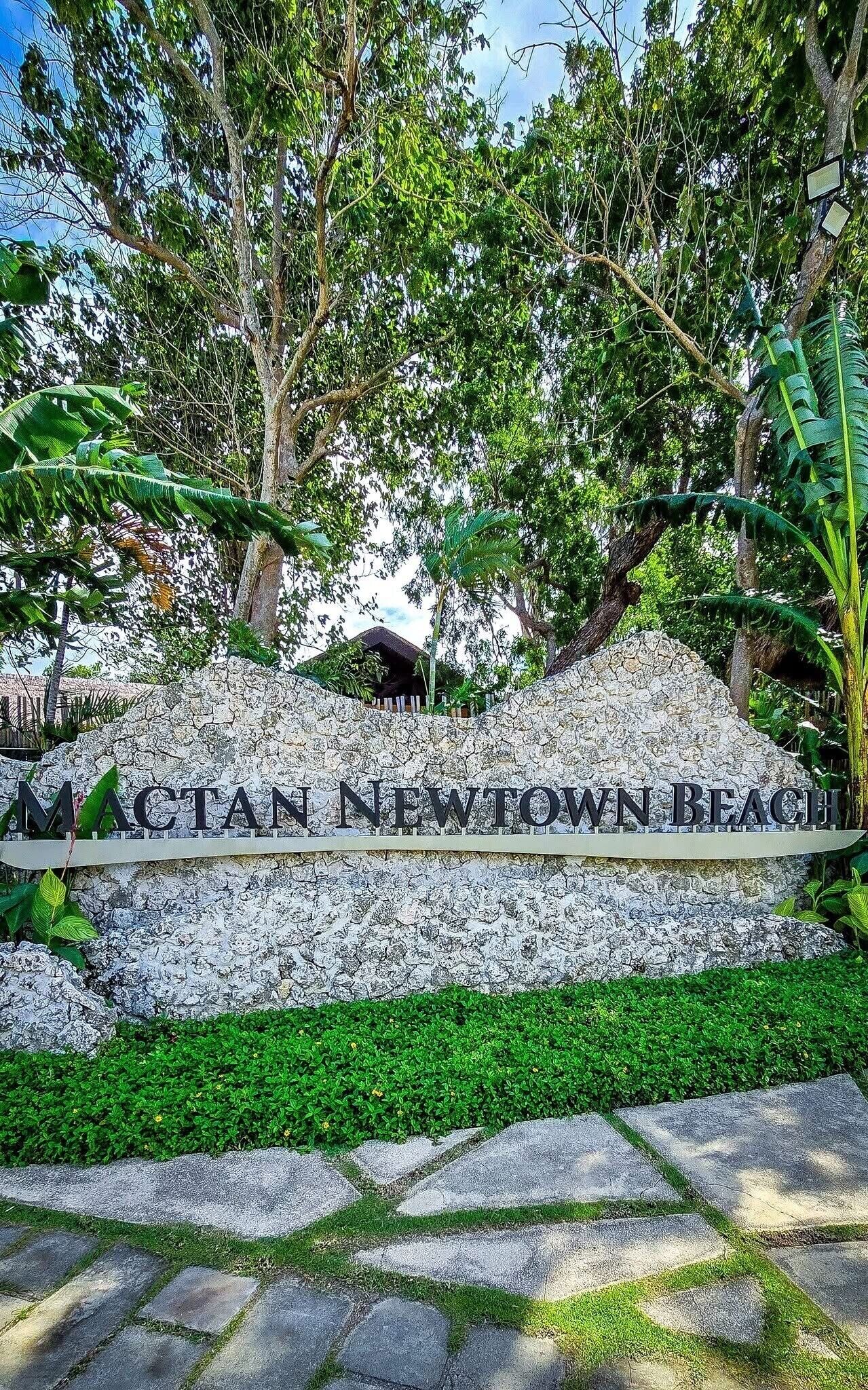 A stone wall with 'MACTAN NEWTOWN BEACH' signage, surrounded by lush greenery and trees.