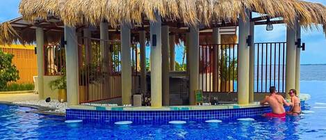 A pool area with a thatched-roof structure, clear blue water, and a view of the sea.