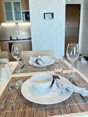 A dining table set with white plates, bowls, and clear glasses, with a kitchen in the background. - Zion Property Mactan Newtown Condo (Lapu-Lapu)