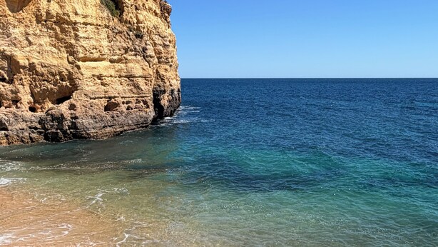 Un paysage côtier avec une eau d’un bleu limpide, une plage de sable et une falaise rocheuse.