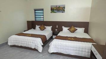 A hotel room with two beds, a nightstand, a bedside table, a window with blinds, and a painting on the wall.
