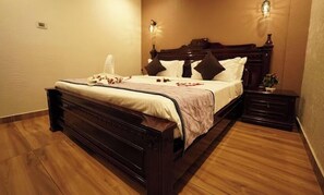 A neatly made bed with a wooden headboard, a bedside table with a lamp, and a small nightstand. - AAR Royal Park (Palayankottai)