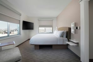 Ein modernes Hotelzimmer mit einem großen Bett, einem Flachbildfernseher, einem Nachttisch, einem kleinen Schreibtisch und einem Fenster mit Stadtblick.