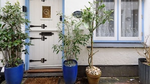 Un bâtiment blanc avec une porte et une fenêtre bleues, des plantes en pot devant, et une enseigne indiquant « POUPÉES ET PETITES MAISONS ».