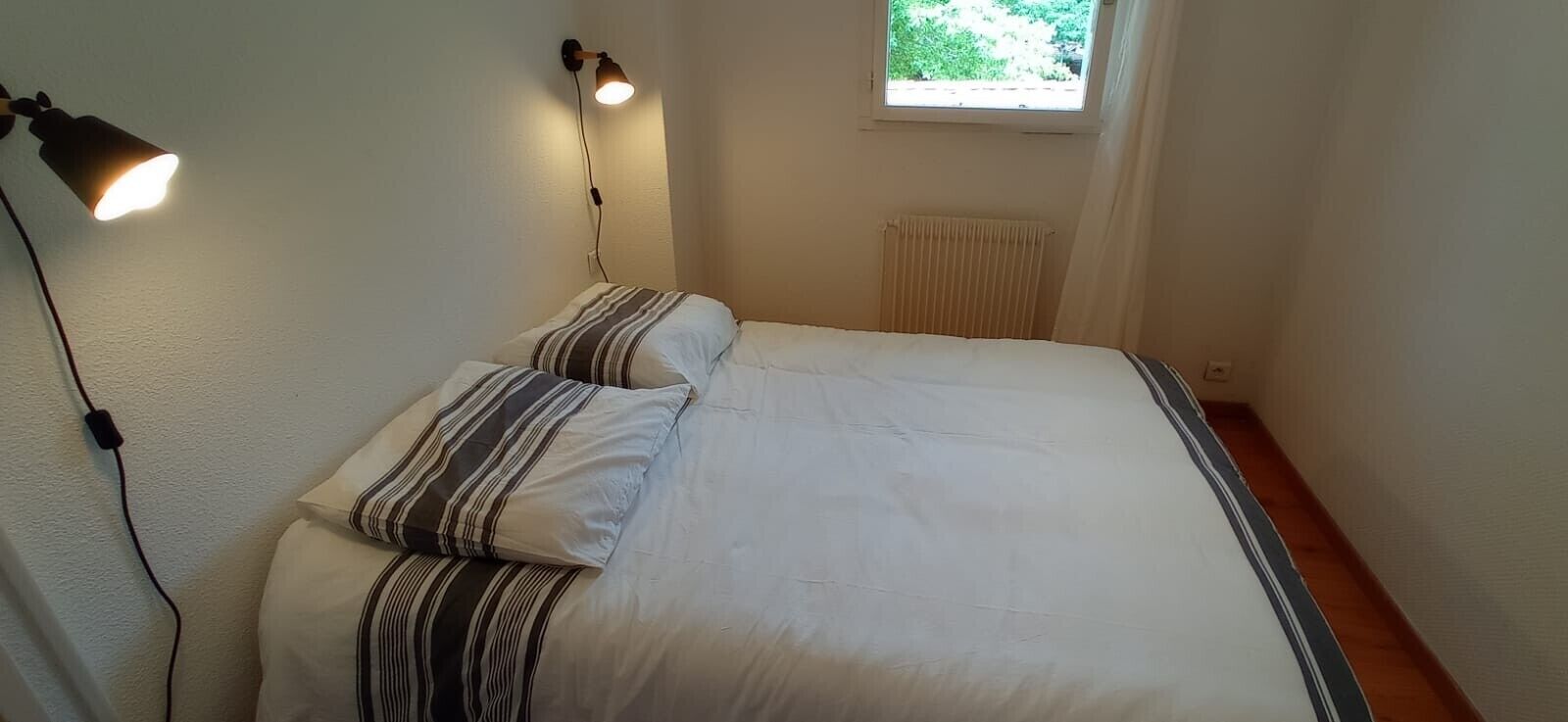 A bedroom with a bed, two bedside lamps, a window with curtains, and a radiator.