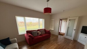 A spacious living room with a red sofa, a beige sofa, and a wooden floor. There is a door leading to another room and a window with blinds. - Carhan River  Holiday Home - Cahersiveen (Cahersiveen)