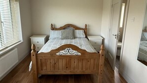 A bedroom with a wooden bed, white bedside tables, a window with blinds, and a door leading to another room. - Carhan River  Holiday Home - Cahersiveen (Cahersiveen)