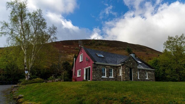 A stone house with a red door and a stone extension, set on a grassy hill with a river in the foreground. - An Charraig - Killarney (Killarney)