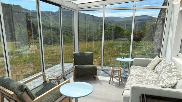 Un invernadero con un sofá, sillas y una mesa, que ofrece vistas a un paisaje montañoso.