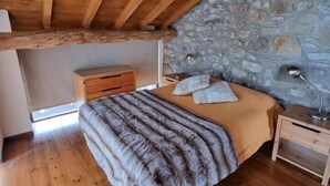 Un dormitorio con techo de madera, pared de piedra, suelo de madera, cama con manta, mesita de noche y lámpara.