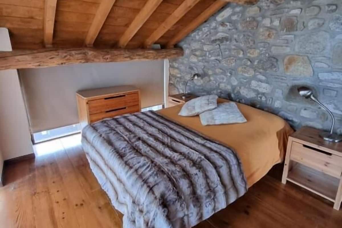 Un dormitorio con techo de madera, pared de piedra, suelo de madera, cama con manta, mesita de noche y lámpara.