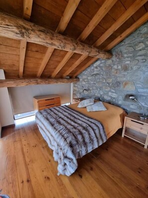 A bedroom with a wooden ceiling, stone wall, wooden floor, bed with a blanket, bedside table, and lamp.
