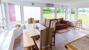 A room with a wooden table, a sofa, and a chair, large windows, and a sign that says 'Be our guest.' - Valentia central,  high spec bungalow wifi &views (Reenglass)