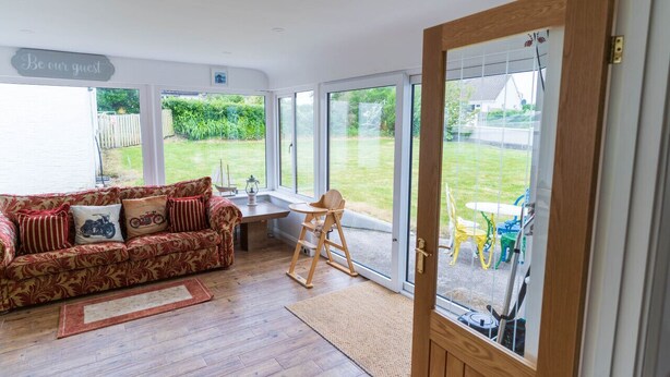 A room with a wooden floor, a sofa with patterned cushions, a wooden chair, and a large sliding glass door leading to an outdoor area with a lawn and chairs. - Valentia central,  high spec bungalow wifi &views (Reenglass)