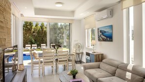 A modern living room with a sofa, a dining table with chairs, a TV, and a painting on the wall. - Milos lifestyle villas H9- 4 bedroom villa with private pool (Paralimni)