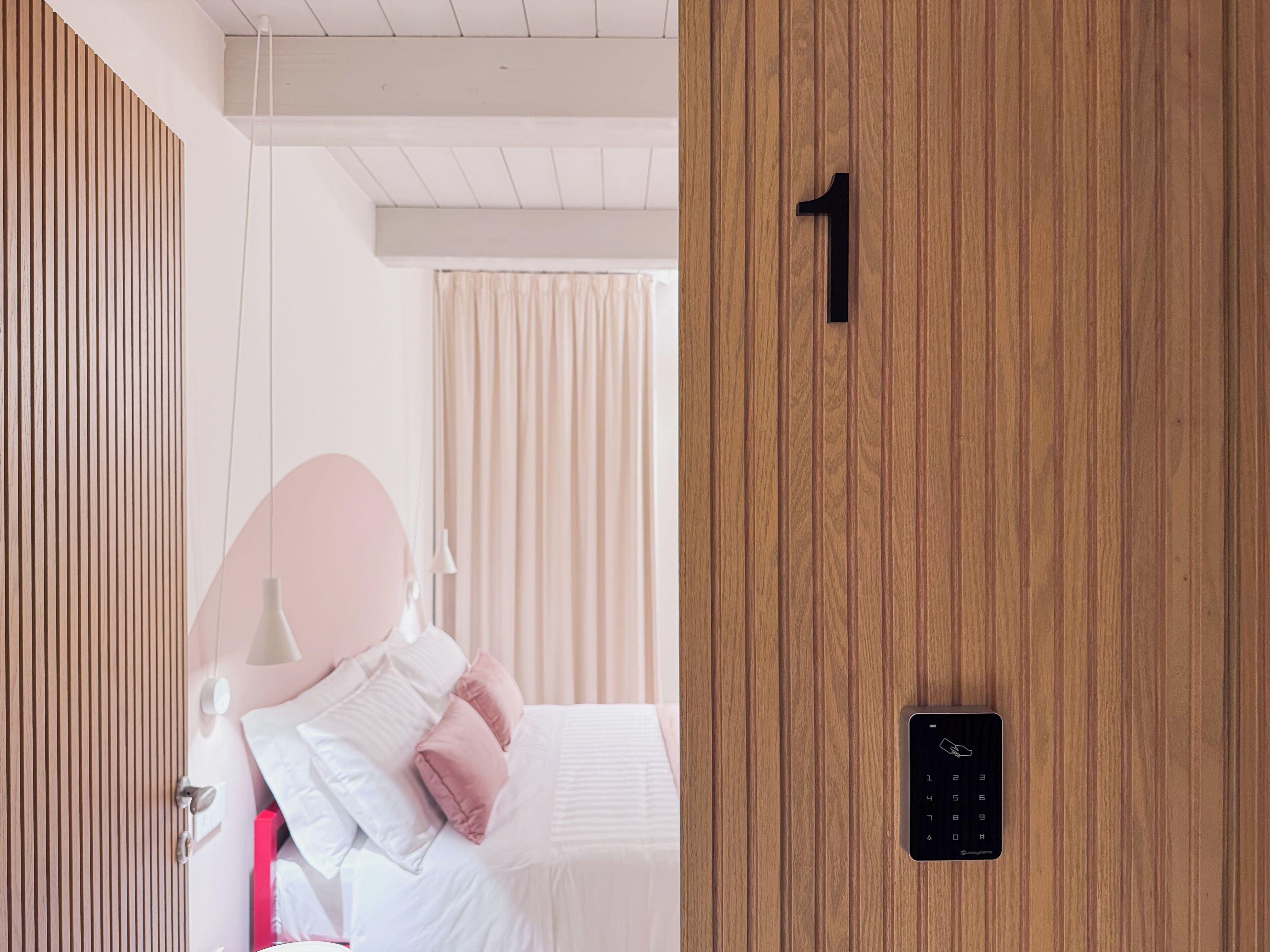 A hotel room with a wooden door marked with the number 1, a bed with white bedding and pink pillows, a bedside table with a lamp, and a window with curtains.