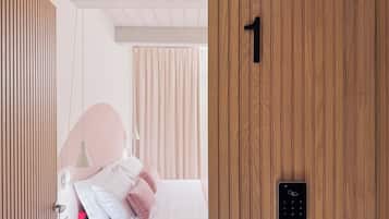 A hotel room with a wooden door marked with the number 1, a bed with white bedding and pink pillows, a bedside table with a lamp, and a window with curtains.