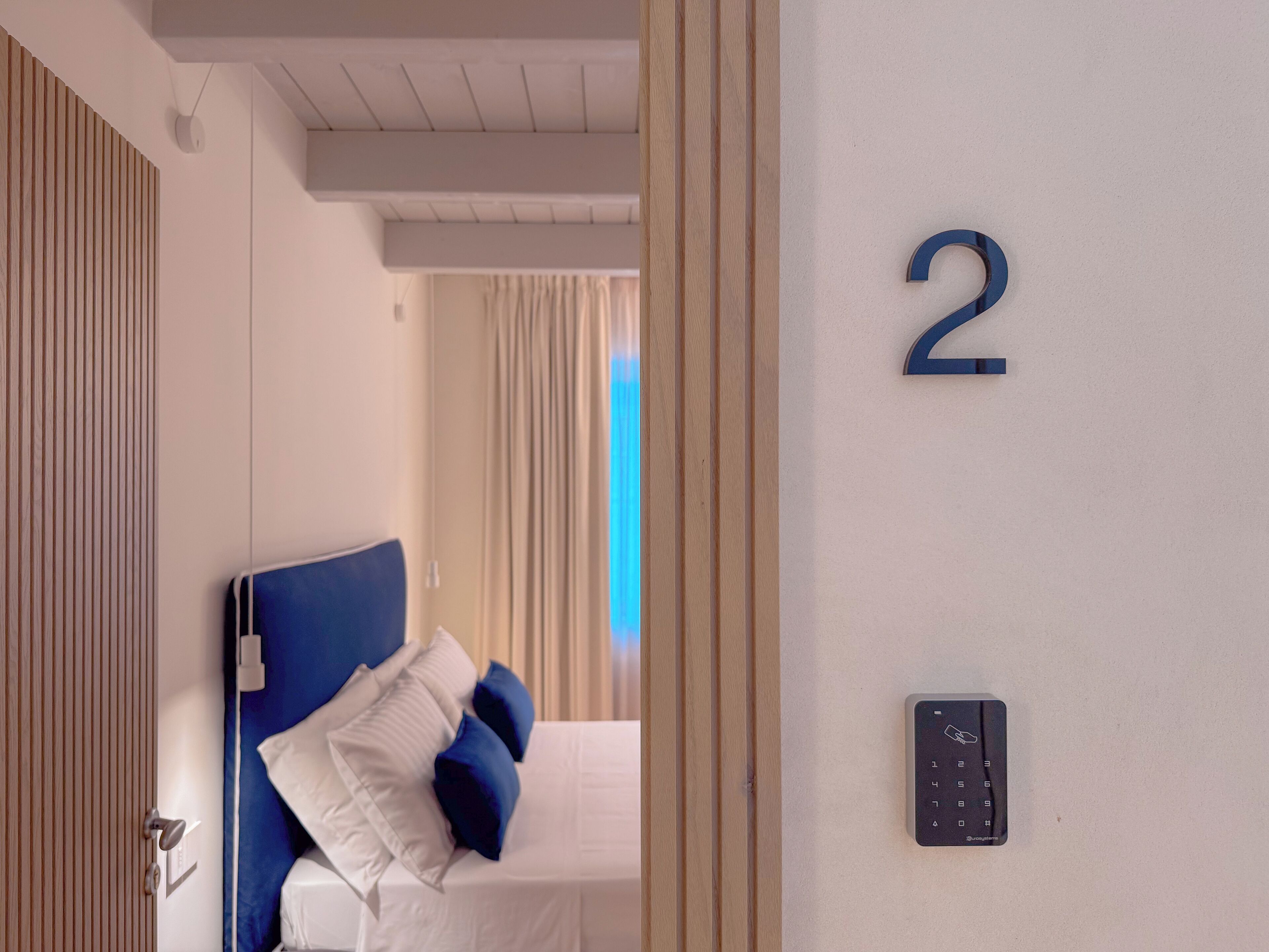 A hotel room with a bed, a wall-mounted number '2', and a door with a wooden panel design.