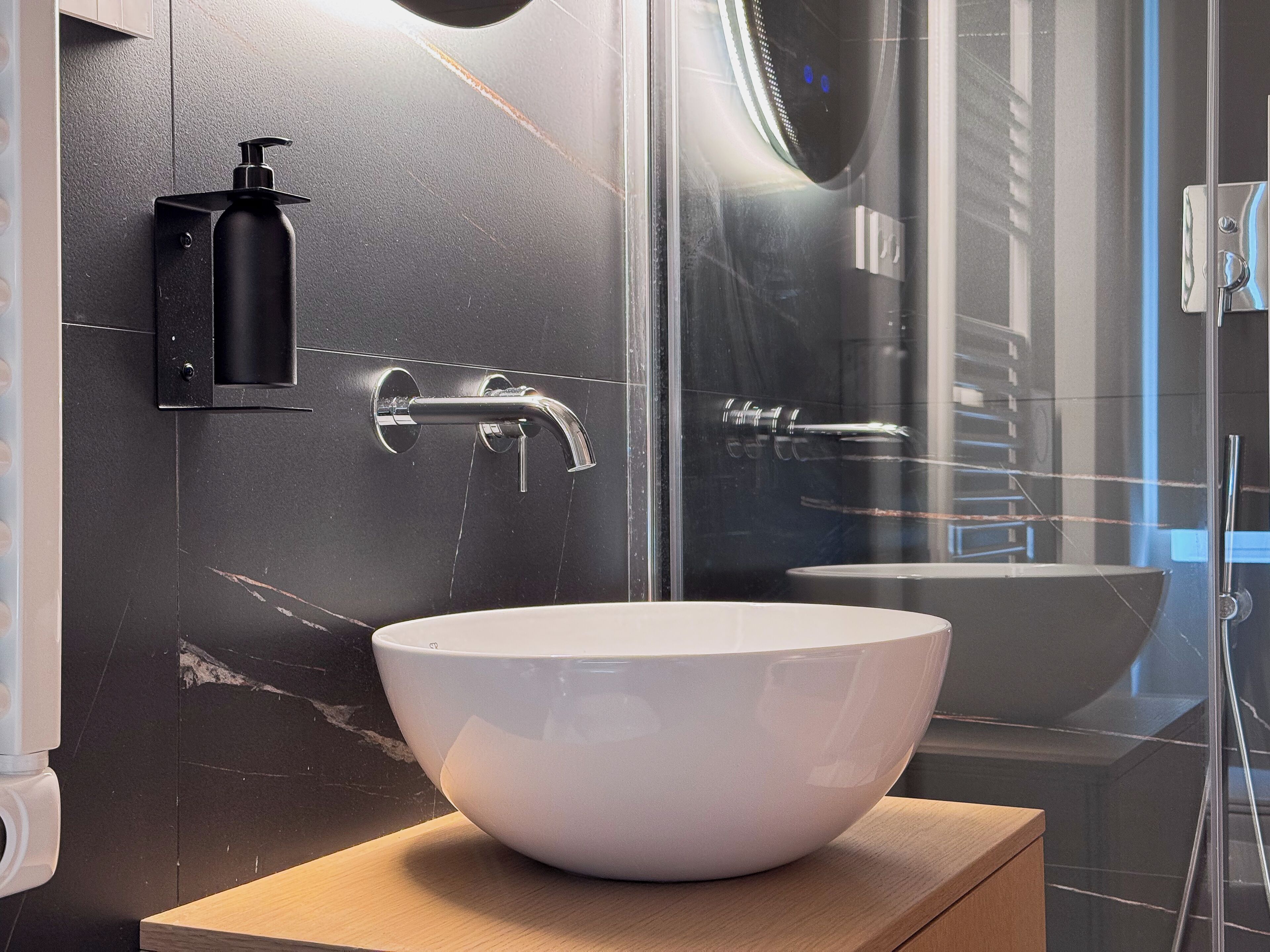 A modern bathroom with a white vessel sink, a black soap dispenser, and a glass shower enclosure.