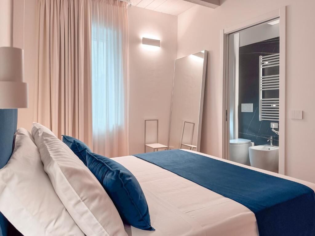 Una camera d'hotel con un letto, una finestra con tende, uno specchio e un bagno visibile attraverso una porta aperta.