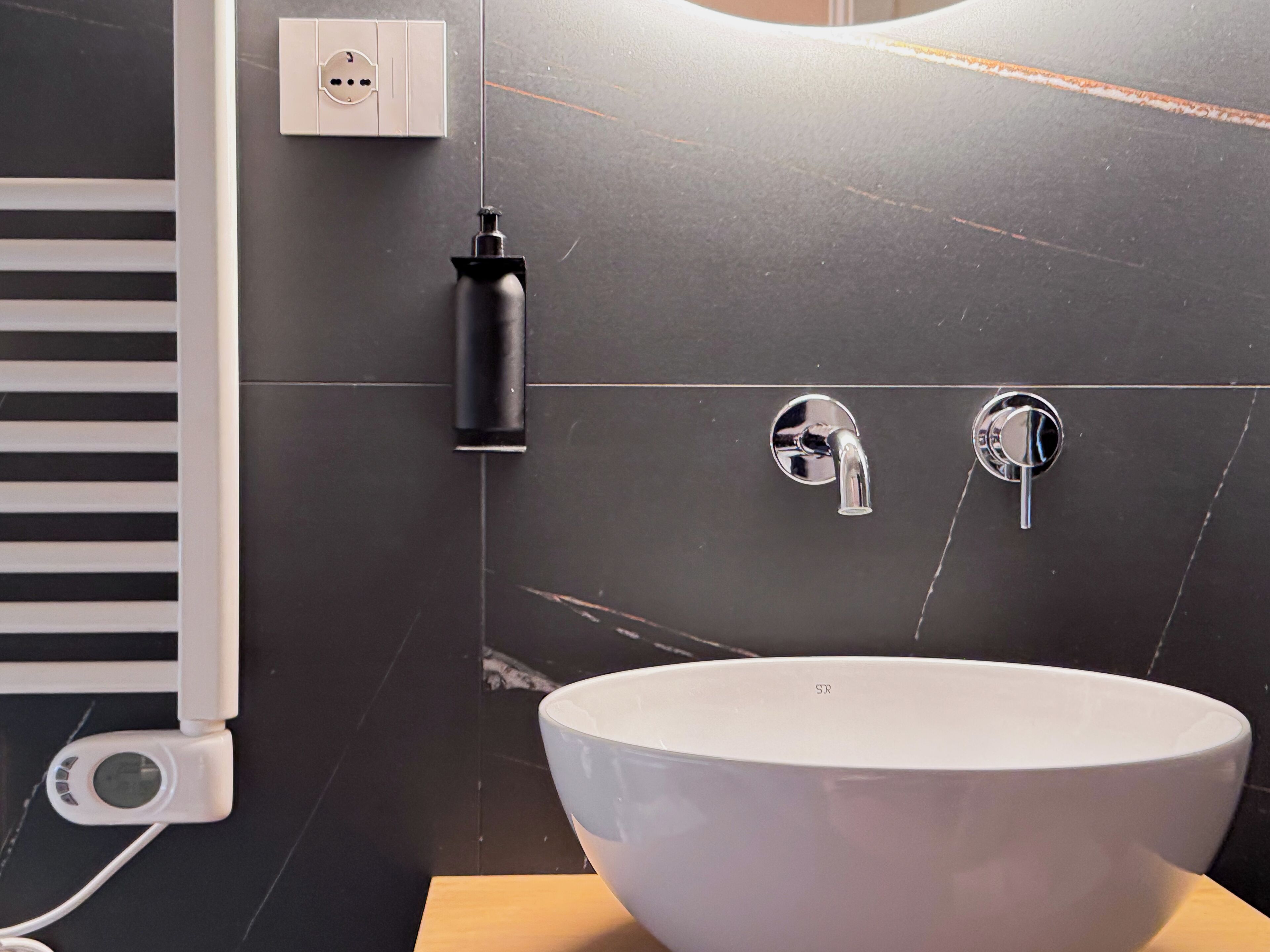 Une salle de bain moderne avec un lavabo blanc, deux robinets chromés, un distributeur de savon noir et une prise électrique blanche fixée au mur.