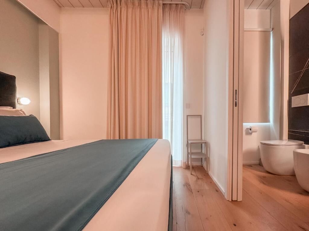 A bedroom with a bed, a bedside table, a window with curtains, a bathroom with a toilet and sink, and a wooden floor.