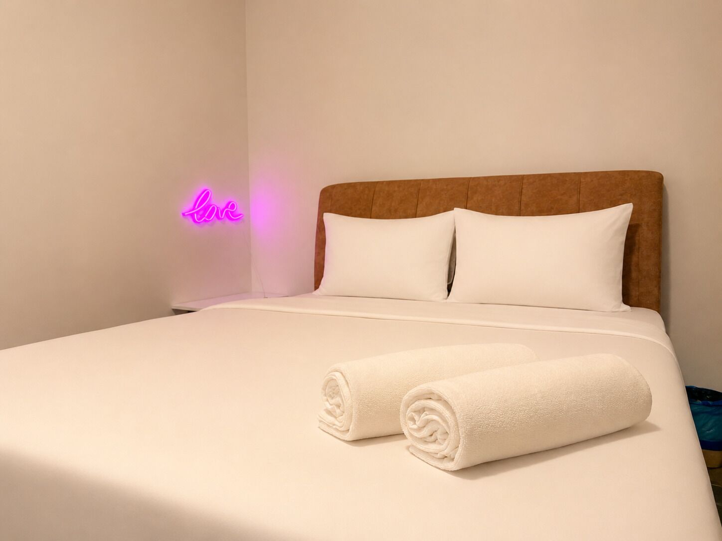 Un lit bien fait, avec deux serviettes enroulées, une tête de lit en bois et un néon accroché au mur.