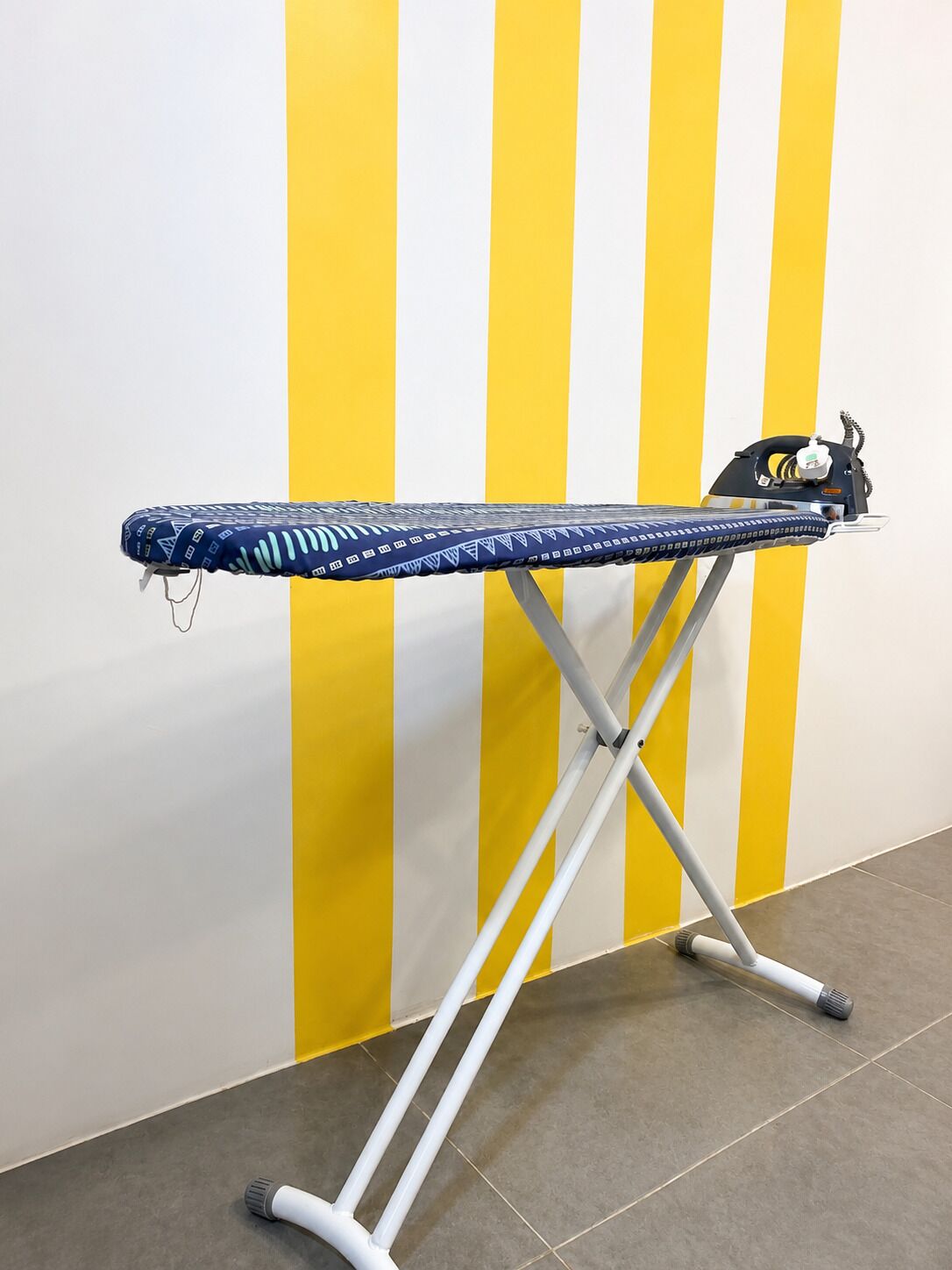 Ein Bügelbrett mit einer blauen, gemusterten Auflage, ein schwarzes Bügeleisen und eine gelb-weiß gestreifte Wand im Hintergrund.