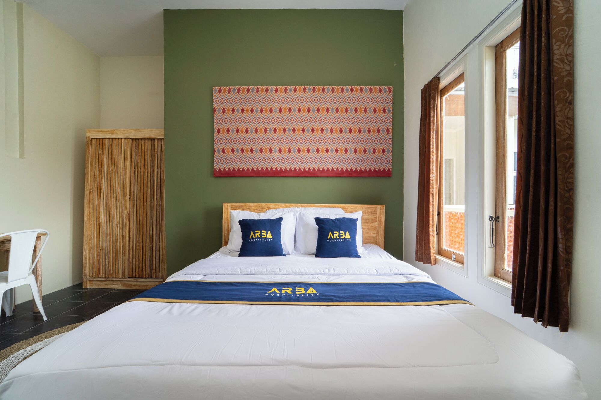 A hotel room with a bed, a wooden wardrobe, a chair, a patterned tapestry, and a window with curtains.
