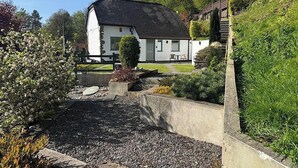 Una casa blanca con techo oscuro, rodeada por un jardín con senderos de grava y diversas plantas.
