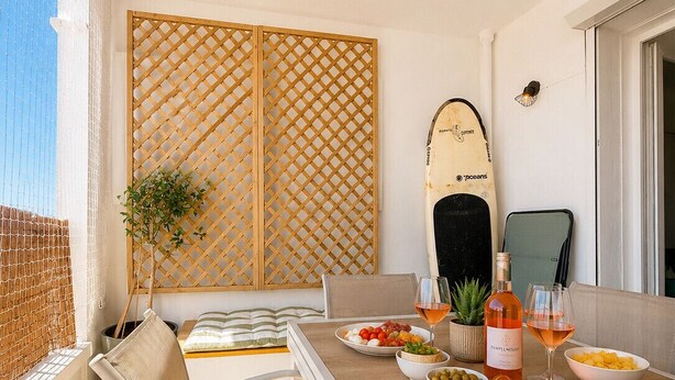 Un balcon ensoleillé avec une table dressée pour le dîner, une planche de surf et une plante en pot.