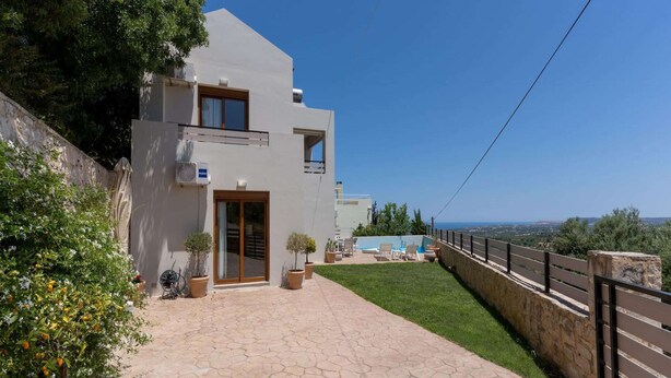 Una casa moderna a due piani con vialetto lastricato, piccolo prato e vista sul mare.
