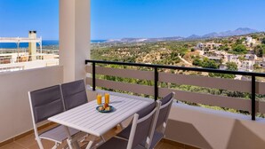 Un balcone con tavolo e sedie, vista su un paesaggio costiero e catene montuose in lontananza.