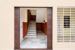 A hallway with a staircase leading to a door, a fire extinguisher on the wall, and a window with a metal grid. - Classic Guwahati Stay Inn (Guwahati)