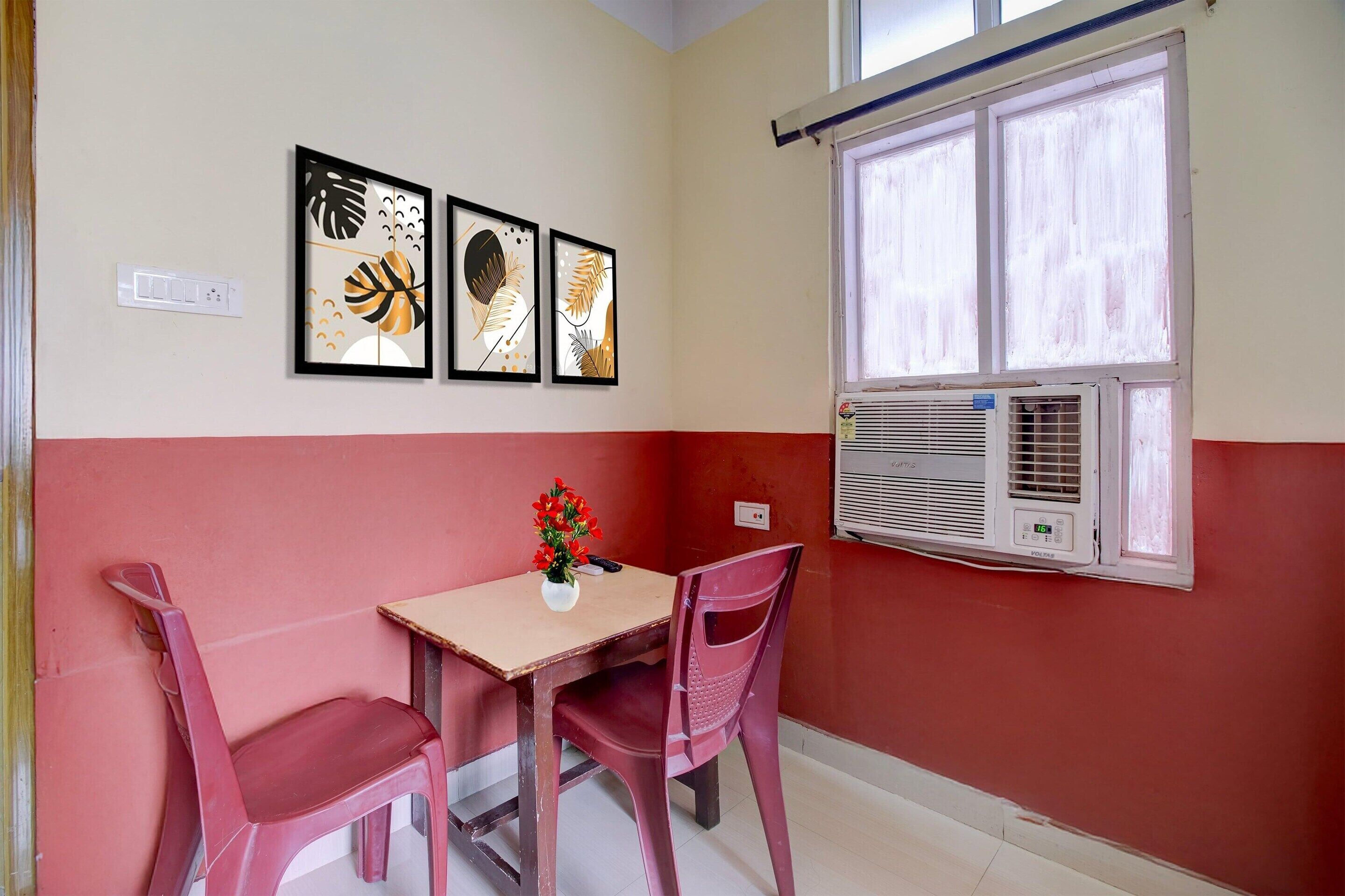 A room with a table, two chairs, a window with an air conditioner, and three framed artworks on the wall.