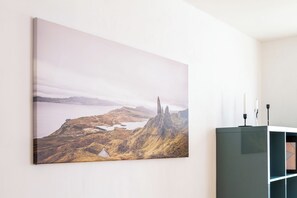 Une peinture de paysage accrochée au mur, un meuble de couleur sombre orné d’un motif géométrique et un mur blanc.