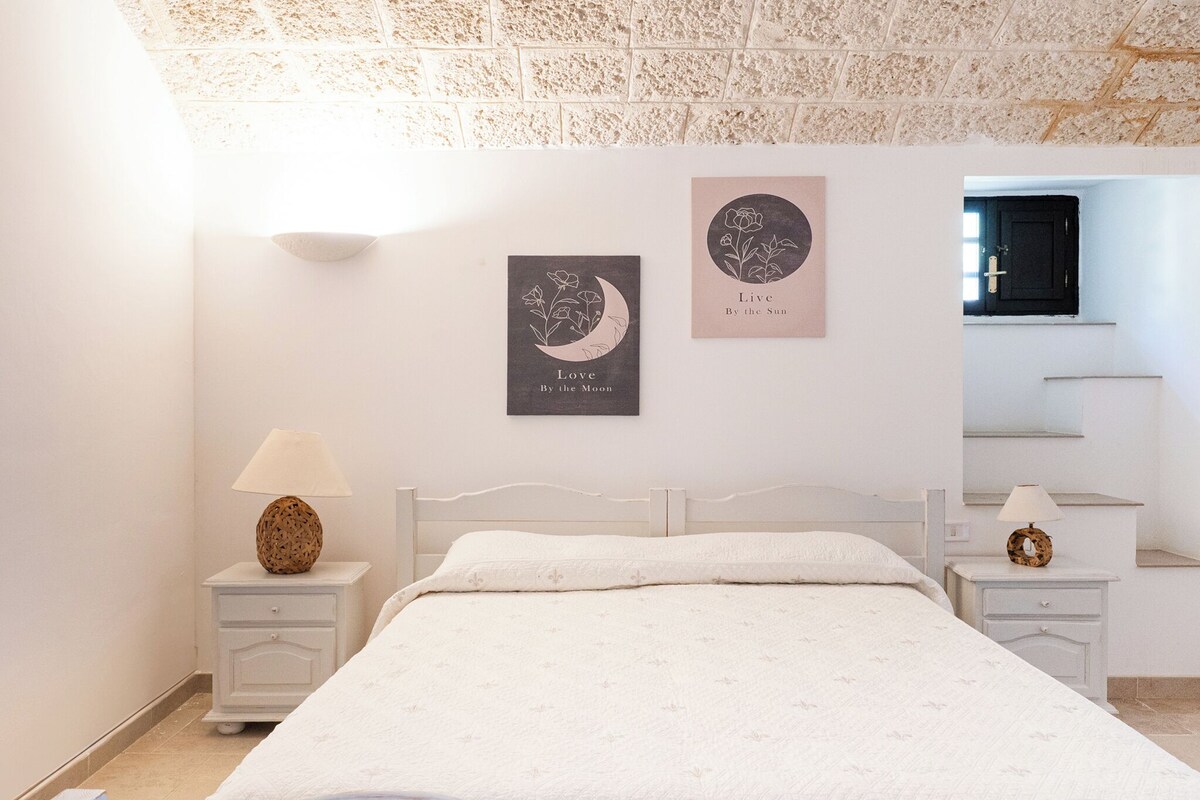 A bedroom with a bed, two nightstands, a wall-mounted lamp, and two framed artworks.
