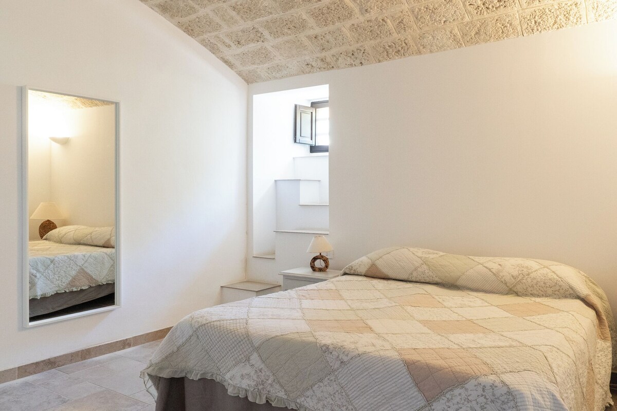 A bedroom with a bed, a bedside table, a mirror, and a window.