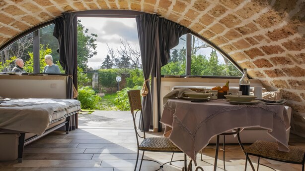 An arched room with a bed, table, and chairs, a window with curtains, and a view of greenery outside.