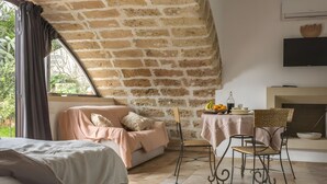 A room with a brick wall, a bed, a sofa, a dining table with chairs, and a television mounted on the wall.