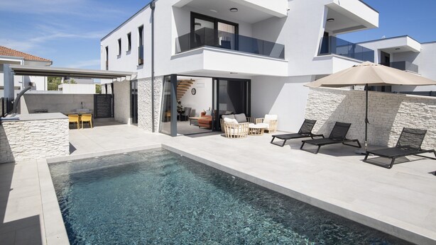 Ein modernes, zweistöckiges Haus mit Swimmingpool, Sitzgelegenheit im Freien und blauem Himmel.