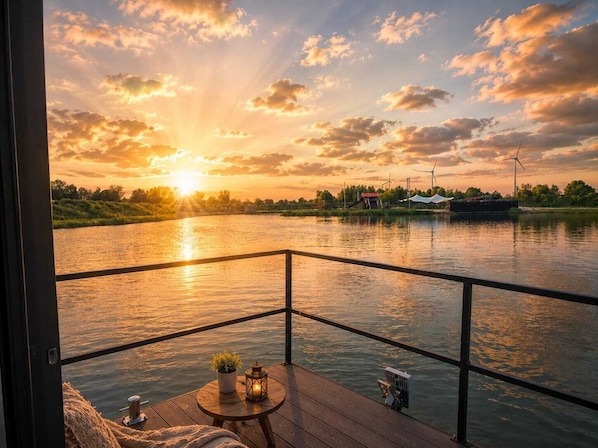 Une terrasse offrant une vue sur une rivière au coucher du soleil, une petite table avec une plante et, au loin, des éoliennes.