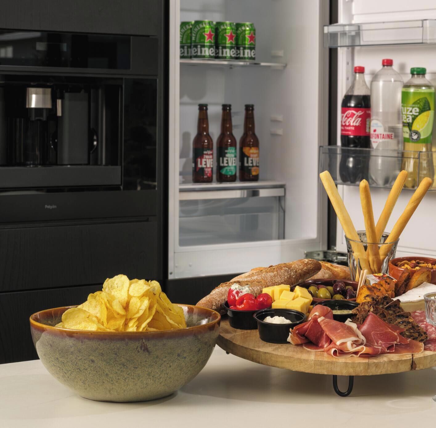 Eine Küche mit einem Kühlschrank voller Getränke, einer Schüssel Chips und einer Platte mit Wurstwaren und Käse.