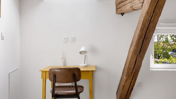 Et værelse med skrivebord, stol, lampe og et vindue med udsigt til grønne områder.