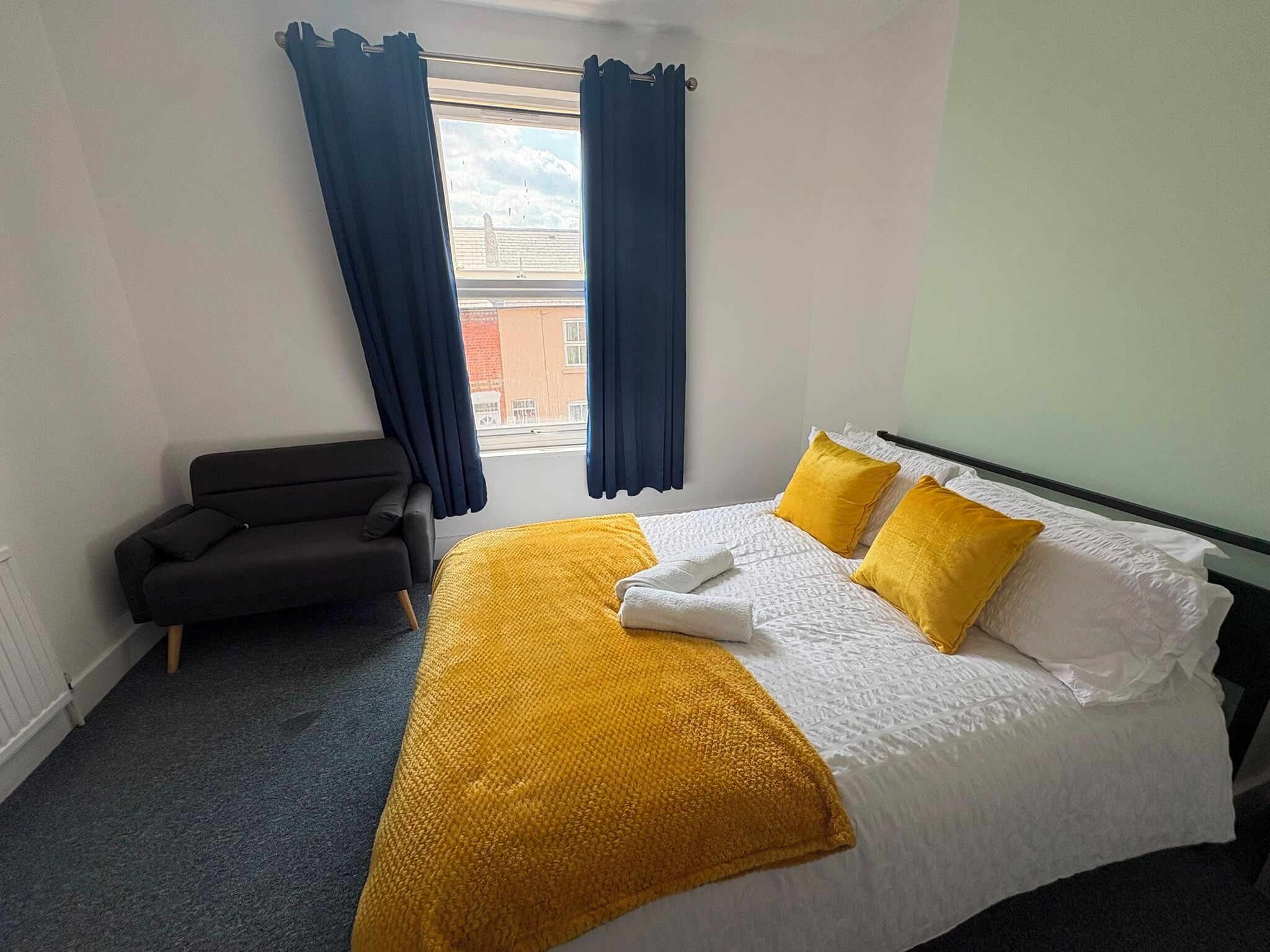 A hotel room with a bed, a chair, a window with curtains, and a view of buildings outside.